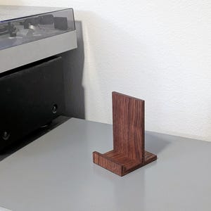 A small, rectangular wooden record stand with a rich, reddish-brown finish. The stand has a vertical back and a horizontal base, designed to hold a vinyl record upright. The wood grain is visible, adding to its natural aesthetic.