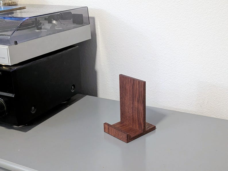 A small, rectangular wooden record stand with a rich, reddish-brown finish. The stand has a vertical back and a horizontal base, designed to hold a vinyl record upright. The wood grain is visible, adding to its natural aesthetic.