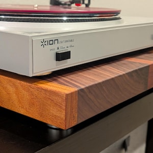 May include: A light gray ION USB turntable with a red vinyl record sits atop a thick, rectangular wooden platform featuring a multi-toned grain pattern. The turntable front panel displays the ION logo and speed settings for 33 and 45 RPM.