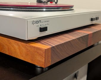 May include: A light gray ION USB turntable with a red vinyl record sits atop a thick, rectangular wooden platform featuring a multi-toned grain pattern. The turntable front panel displays the ION logo and speed settings for 33 and 45 RPM.