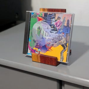 A CD in a clear case is displayed in a wooden stand. The CD cover features a colorful abstract design with the word "Dreams" in a stylized font. The stand is made of layered wood.