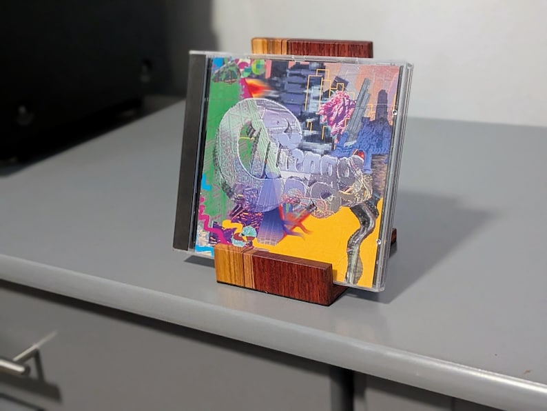 A CD in a clear case is displayed in a wooden stand. The CD cover features a colorful abstract design with the word "Dreams" in a stylized font. The stand is made of layered wood.