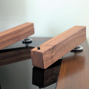 以下が含まれることがあります： 濃い茶色の仕上げが施された2つの長方形の木製オブジェクト。それぞれ銀色のベースに取り付けられています。各木片の上部には小さな黒いネジが見えます。オブジェクトは光沢のある暗い表面に配置されています。