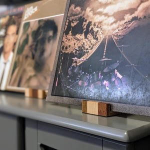 Close-up view of several vinyl records displayed on a gray surface using wooden record stands. The records feature various album art, including a landscape scene with a road and a drum set, and portraits. The stands are made of light and dark brown wood, providing a rustic contrast to the gray surface.