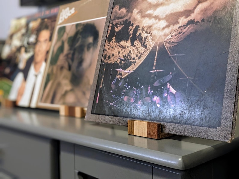 Close-up view of several vinyl records displayed on a gray surface using wooden record stands. The records feature various album art, including a landscape scene with a road and a drum set, and portraits. The stands are made of light and dark brown wood, providing a rustic contrast to the gray surface.