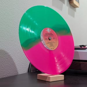 May include: A split-color vinyl record featuring a vibrant green top half and a bright pink bottom half. The center label displays the text Tom Petty and the Heartbreakers, Live at the Fillmore 1997, and a tracklist. The record rests on a wooden stand.