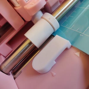 Puede incluir: Primer plano de una máquina de manualidades rosa y blanca con una varilla de metal plateado. La máquina tiene una esterilla de corte azul claro con marcas de medición blancas. Los componentes son de plástico.