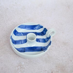 Può includere: Un portacandele in ceramica bianca con un motivo a righe blu. Il portacandele ha una base circolare, una coppa centrale per la candela e un piccolo manico. Le strisce blu sono dipinte a mano, conferendogli un aspetto artigianale unico.