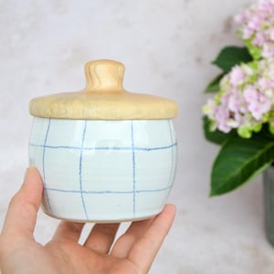 Könnte beinhalten: Ein kleines, rundes Keramikgefäß mit weißer Glasur und blauem Karomuster. Das Gefäß hat einen glatten, hellen Holzdeckel mit einem mittigen Knaufgriff. Der Behälter besitzt eine zylindrische Form mit leicht gewölbten Seiten und einem flachen Boden.
