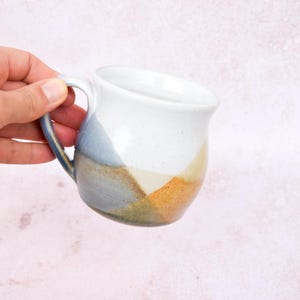 Könnte beinhalten: Ein Keramikbecher mit rundem Körper und geschwungenem Henkel. Der Becher hat eine gesprenkelte weiße Glasur im oberen Bereich, die im unteren Teil in geometrische Farbblöcke aus gedämpftem Blau, sanftem Gelb und erdigem Orange übergeht.