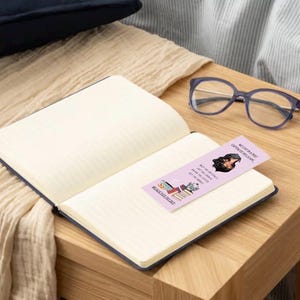 May include: A pink bookmark with the text "We can be a bit dramatic, can't we?" and a cartoon image of a woman with a book. The bookmark is on top of a closed notebook with a white background.