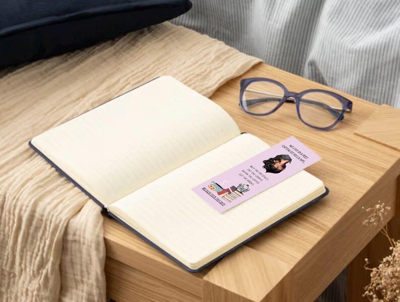 May include: A pink bookmark with the text "We can be a bit dramatic, can't we?" and a cartoon image of a woman with a book. The bookmark is on top of a closed notebook with a white background.