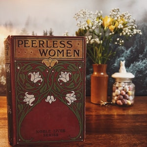 May include: An antique book, "Peerless Women", with a decorative cover in red, green, and white floral patterns. A vase of yellow flowers, a small brown jar, and a glass jar with sweets are placed beside the book on a wooden surface.