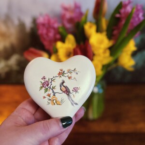 May include: An off-white, heart-shaped trinket box with a bird and floral design. The box has a colourful illustration of a bird on a branch with flowers. A blurred bouquet of flowers is in the background.