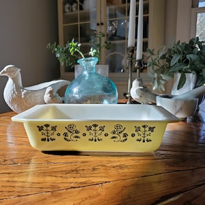 Puede incluir: Una fuente rectangular amarilla para hornear con un patrón floral negro. La fuente está sobre una mesa de madera con un pájaro de cerámica blanco y un jarrón de vidrio azul en el fondo.