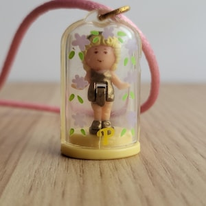 May include: A vintage miniature doll pendant necklace. The doll is inside a clear plastic case decorated with purple flowers and green leaves. The doll has blonde hair and is wearing a gold-colored dress. The necklace has a pink cord.