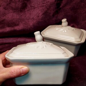 May include: Two white ceramic rectangular lidded dishes. The lids have a floral design and a small knob. One dish is held by a hand. The dishes are on a dark burgundy surface.