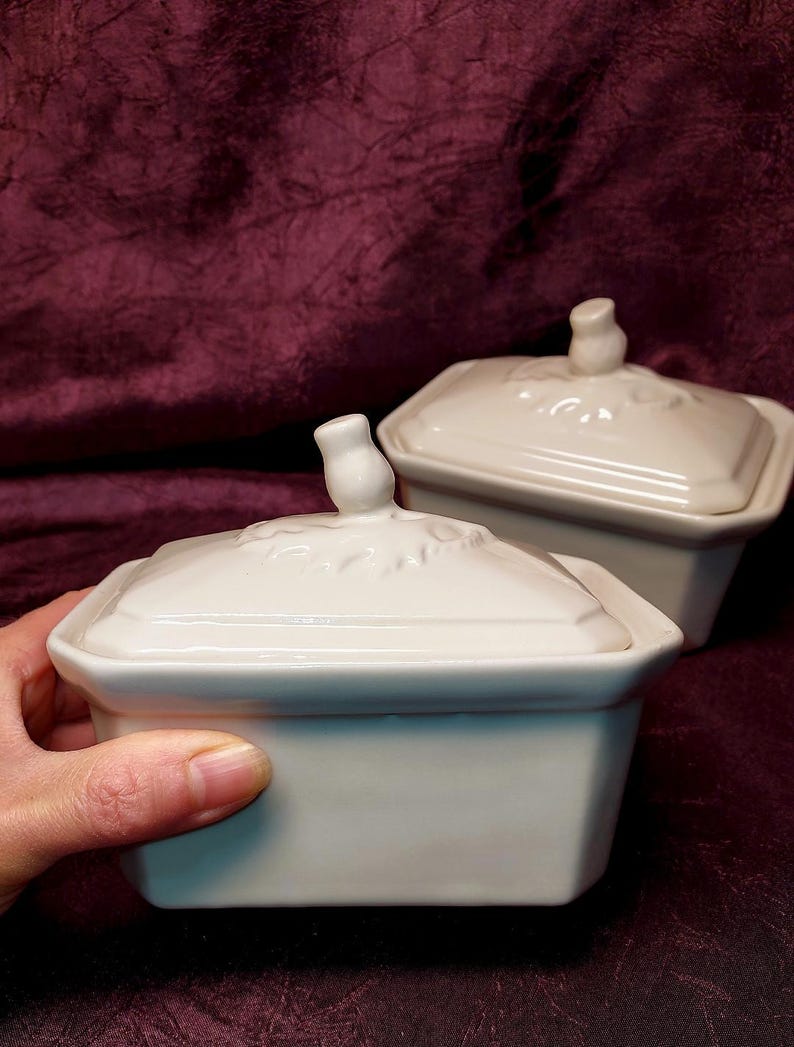 May include: Two white ceramic rectangular lidded dishes. The lids have a floral design and a small knob. One dish is held by a hand. The dishes are on a dark burgundy surface.