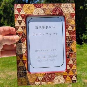 Puede incluir: Un marco de fotos rectangular con un patrón geométrico de incrustaciones de madera Yosegi-zaiku en tonos rojo, amarillo y marrón. El inserto central muestra el texto Hakone Yosegi Zaiku, photo frame y caracteres japoneses sobre el tamaño.