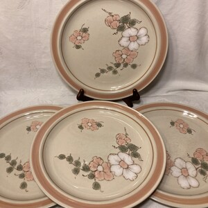 May include: Set of four ceramic dinner plates with a floral design. The plates are beige with a pink rim and feature a delicate floral pattern in shades of pink, white, and green.