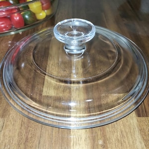 Può includere: Un coperchio in vetro trasparente di forma rotonda con una piccola maniglia circolare. Il coperchio ha un bordo rialzato ed è progettato per adattarsi a una pentola o padella. Il coperchio è su una superficie di legno.