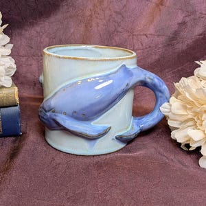 May include: A ceramic mug with a light blue glaze featuring a sculpted, three-dimensional blue whale wrapped around the exterior. The whale's tail forms the handle of the mug, and the body is detailed with small, raised dots along the side.