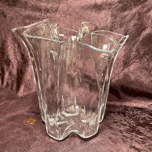 May include: A clear glass vase with a unique, organic shape. The vase has a textured surface and a wide opening. It is made by the brand Iittala.