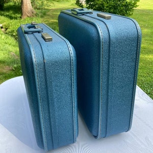 Puede incluir: Dos maletas vintage azules con herrajes plateados. Las maletas están hechas de un material texturizado y tienen un aspecto retro.