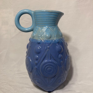 May include: A blue ceramic pitcher with a textured design. The pitcher has a light blue top and a darker blue bottom. The design features circles and triangles.