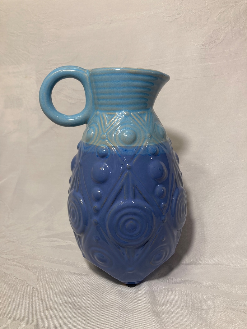 May include: A blue ceramic pitcher with a textured design. The pitcher has a light blue top and a darker blue bottom. The design features circles and triangles.