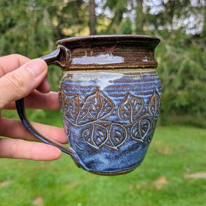 May include: A ceramic mug featuring a dark brown glazed rim and a textured blue body decorated with a repeating embossed leaf pattern. The mug has a curved handle and a wide opening, showcasing a handcrafted finish with visible glaze variations.