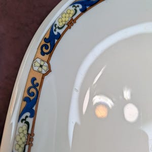 May include: A white ceramic plate featuring a decorative border with a tan band, blue scrollwork, and small yellow floral accents. The rim is accented with geometric shapes in white and green, creating a repeating pattern around the edge.