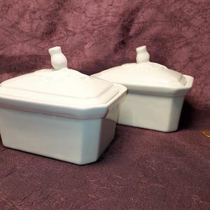 May include: Two white ceramic rectangular lidded dishes. Each dish has a small, decorative handle on the lid. The dishes are on a dark purple surface. A penny is visible in the lower right corner.