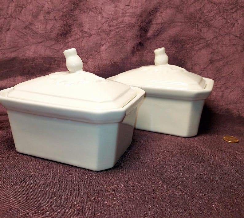 May include: Two white ceramic rectangular lidded dishes. Each dish has a small, decorative handle on the lid. The dishes are on a dark purple surface. A penny is visible in the lower right corner.
