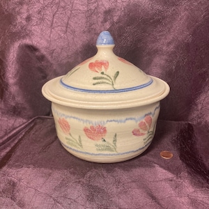 May include: A white ceramic casserole dish with a lid. The dish is decorated with a floral pattern in shades of pink, blue, and green.