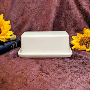 May include: A rectangular white ceramic butter dish with a smooth, solid-colored lid and a matching base. The butter dish is positioned between two bright yellow sunflowers and a dark blue book with gold lettering on the spine.