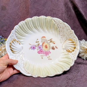 Könnte beinhalten: Eine runde Keramikschale mit gewelltem Rand und einer cremefarbenen, gerippten muschelartigen Textur. Die Mitte zeigt einen Blumenstrauß mit rosa, violetten und gelben Blüten. Zwei goldfarbene Muschelornamente akzentuieren den Schalenrand.