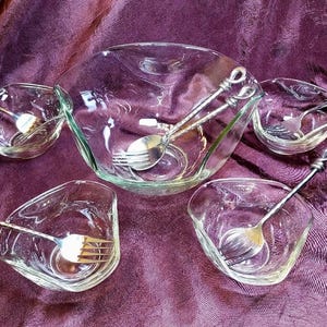 Może przedstawiać: Przezroczysty zestaw do serwowania szkła z dużą miską i czterema mniejszymi miseczkami, każda ze srebrnym przyborem do serwowania. Miski mają unikalny, kanciasty kształt. Zestaw jest ułożony na fioletowym tle z tkaniny.