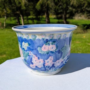 Puede incluir: Una maceta de cerámica con base blanca y un fondo pintado a mano en tono azul. El exterior está decorado con flores rosas y hojas verdes, acentuado por un borde a rayas azules y blancas y un patrón festoneado que se repite.