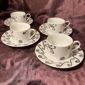 May include: Set of four white teacups and saucers with black swirling designs. The cups and saucers have a simple, elegant design.