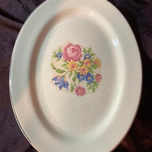 Pode incluir: Um prato oval de cerâmica branca com uma borda dourada e um desenho floral no centro. O desenho apresenta uma rosa rosa com flores amarelas, azuis e rosas que a rodeiam.