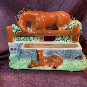 Könnte beinhalten: Ein Keramik-Pflanztopf in Form eines Pferdes und Fohlens. Das Pferd ist braun mit schwarzer Mähne und Schweif und lehnt sich über einen Zaun. Das Fohlen liegt auf dem Boden vor dem Zaun. Der Topf ist mit grünem Gras und Blättern verziert.