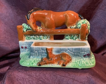 May include: A ceramic planter shaped like a horse and foal. The horse is brown with a black mane and tail, and is leaning over a fence. The foal is lying on the ground in front of the fence. The planter is decorated with green grass and leaves.