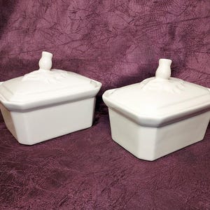 May include: Two white ceramic lidded boxes with a rectangular shape. The lids have a decorative floral design and a small knob. The boxes are sitting on a dark purple surface.