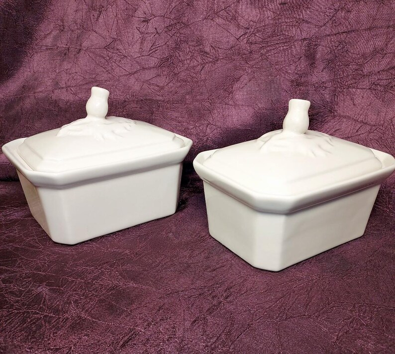 May include: Two white ceramic lidded boxes with a rectangular shape. The lids have a decorative floral design and a small knob. The boxes are sitting on a dark purple surface.