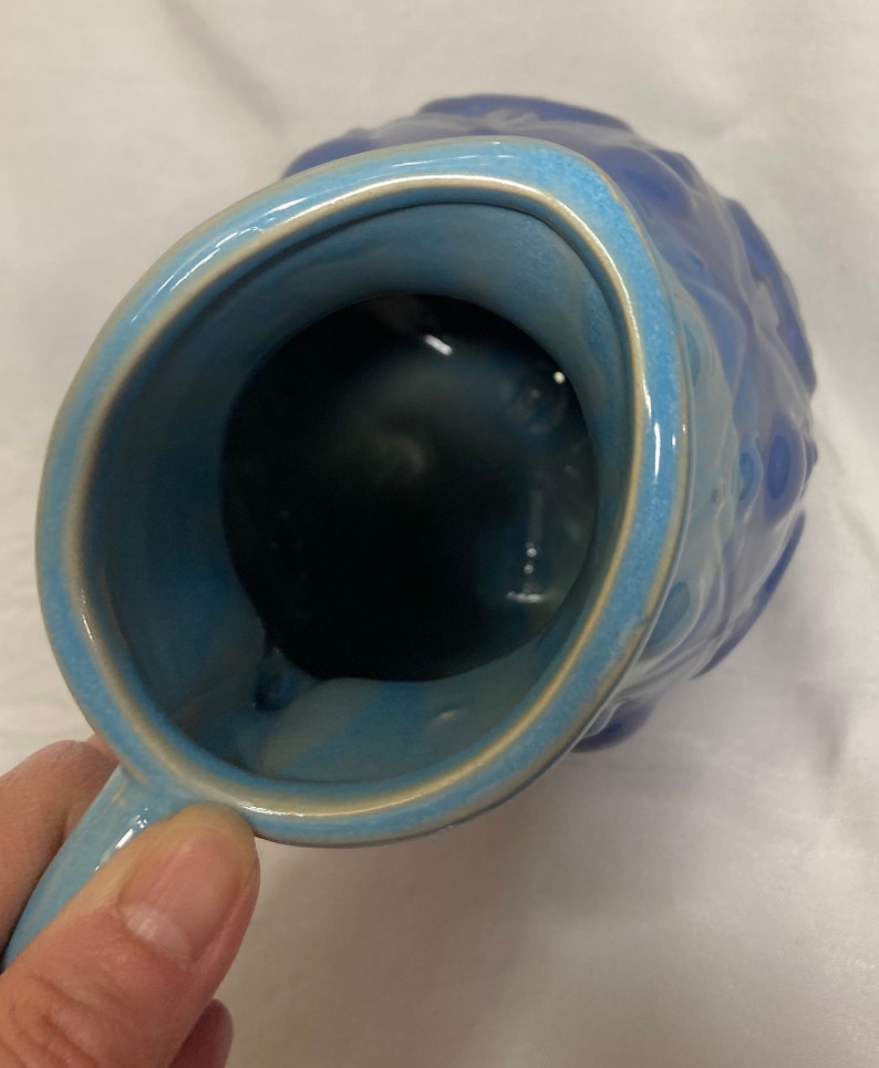 May include: A blue ceramic pitcher with a textured exterior and a smooth, glazed interior. The pitcher has a wide mouth and a handle.