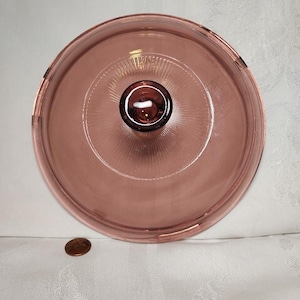 Puede incluir: Una tapa de vidrio rosa con un pomo en la parte superior. La tapa tiene una superficie texturizada y un borde de vidrio transparente.