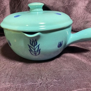 Op de afbeelding: Een turquoise blauwe keramische ovenschaal met een deksel en een handvat. De schaal heeft een blauw en wit tulpenmotief.