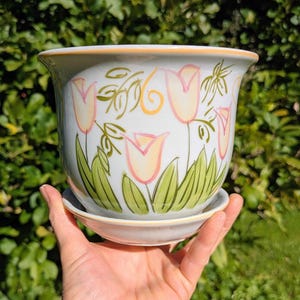 May include: A white ceramic flower pot with a matching saucer, featuring a hand-painted design of pink and yellow tulips with green leaves and stems. The rim of the pot and the saucer are accented with a thin orange line, creating a decorative border.