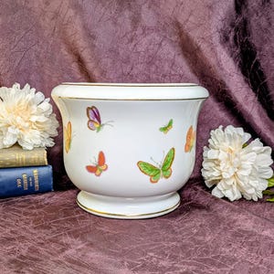 May include: A white ceramic planter with a flared rim and base, featuring a gold-tone trim along the edges. The exterior is decorated with colorful butterfly illustrations in shades of green, orange, yellow, and purple scattered across the surface.